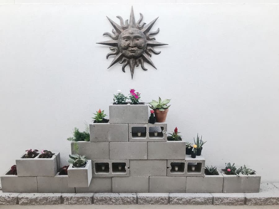 Tiered DIY cinder block planter wall with succulents and flowers, set against a white wall with a decorative sun sculpture for a modern garden display