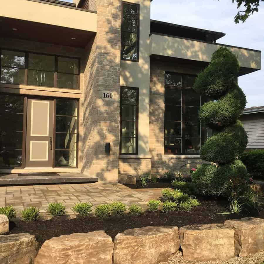 Modern front yard with a spiral topiary, stone landscaping, paved walkway, and a contemporary home with large windows