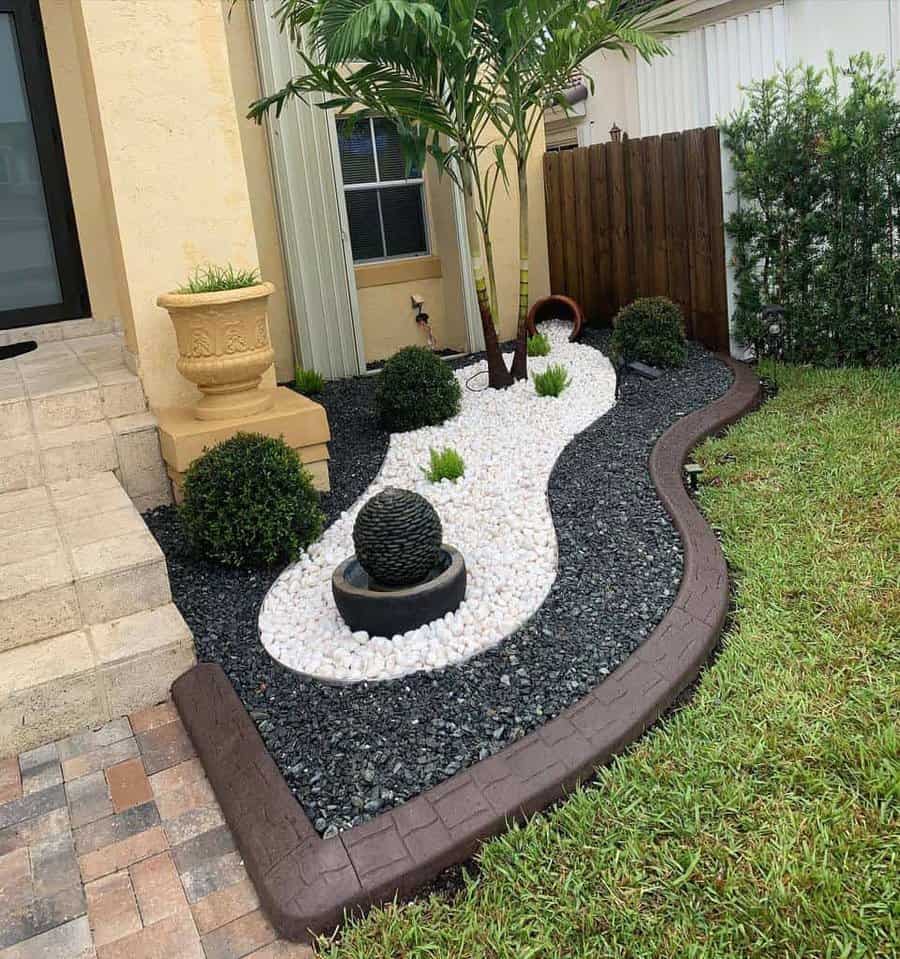 Small front yard landscaping with a tropical tree, decorative white and black stones, curved edging, and a potted plant accent