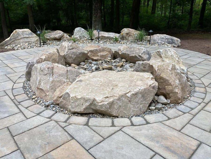 Rustic fire pit surrounded by large boulders, decorative gravel, and a circular stone patio in a wooded outdoor setting