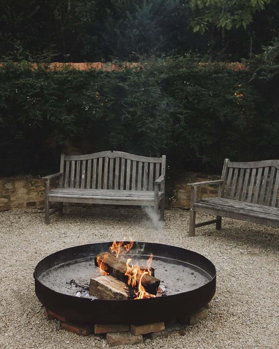 A fire pit with burning logs on gravel, surrounded by two wooden benches, with dense green foliage in the background