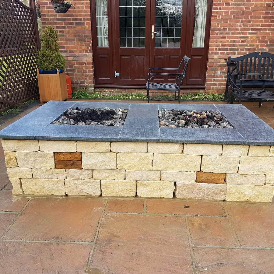 Stone fire pit with black gravel on patio, wooden bench and potted plant nearby, brick house with glass doors in the background