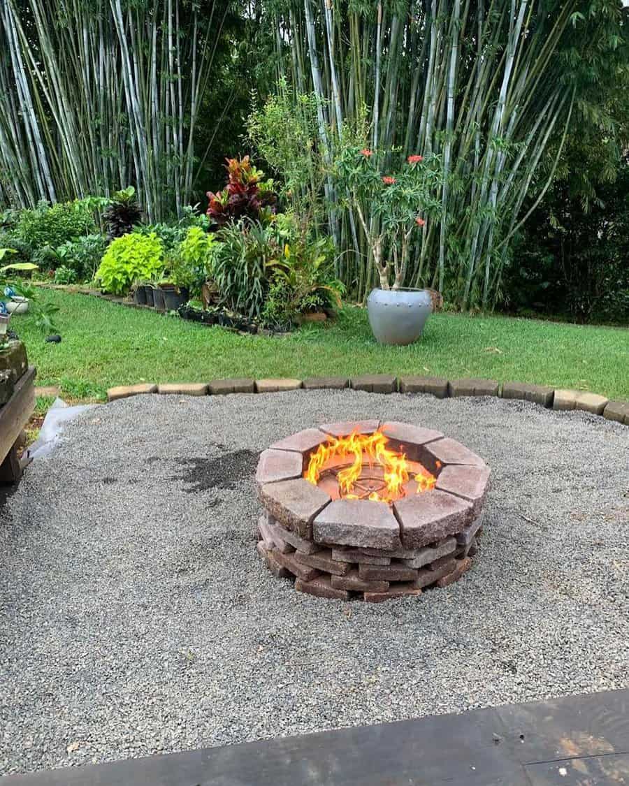 A stone fire pit with bright flames, surrounded by gravel, set in a garden with lush green plants and tall bamboo in the background