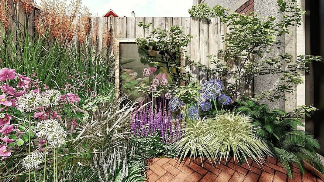 A lush garden with colorful flowers, tall grasses, and green shrubs beside a wooden fence on a brick patio