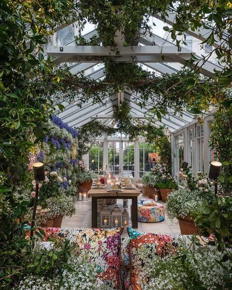 A lush greenhouse with a glass roof, vibrant floral cushions, hanging plants, and lanterns on a table creating a cozy atmosphere