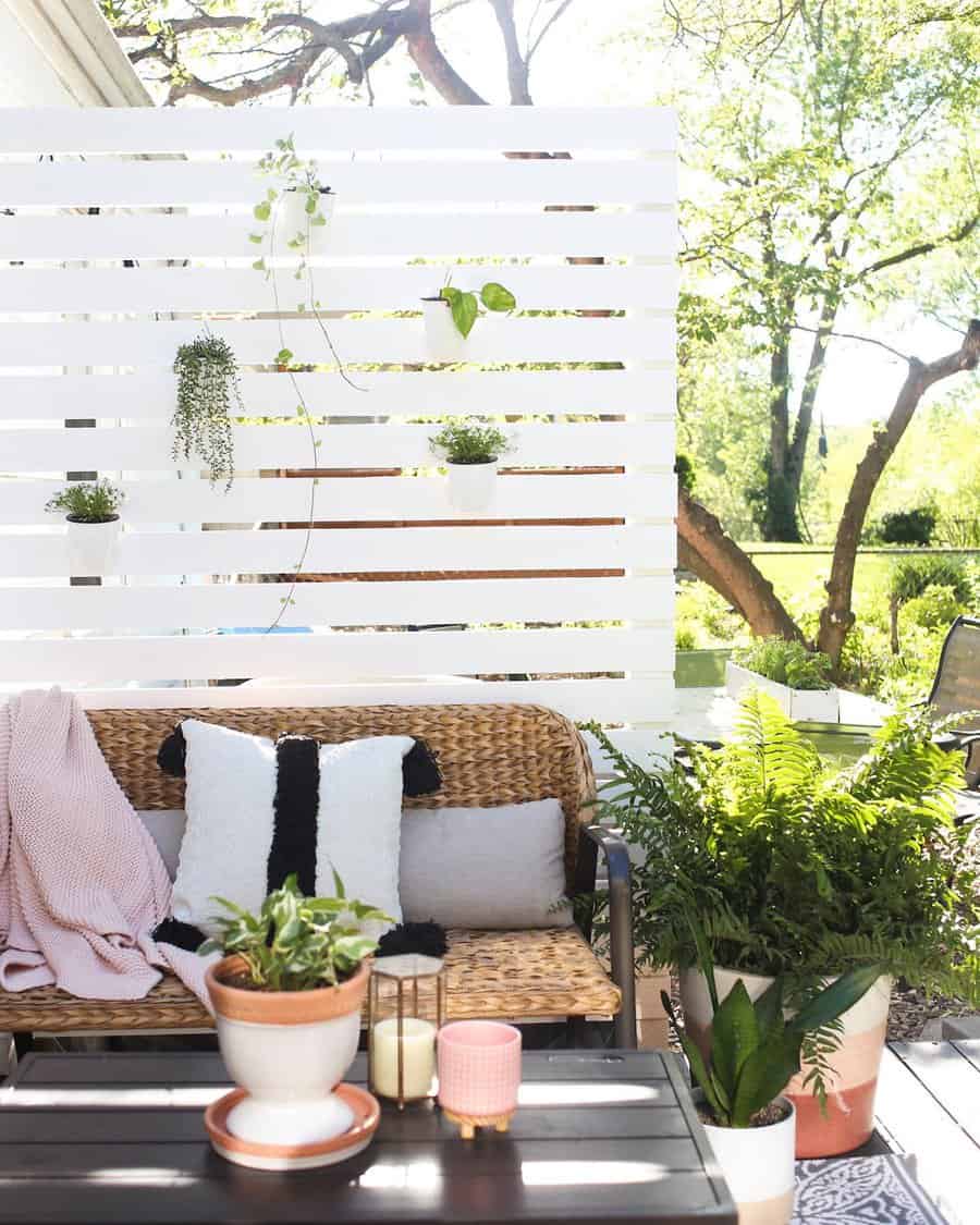 Sunny patio with wicker sofa, cushions, potted plants, and a wooden privacy screen adorned with hanging greenery