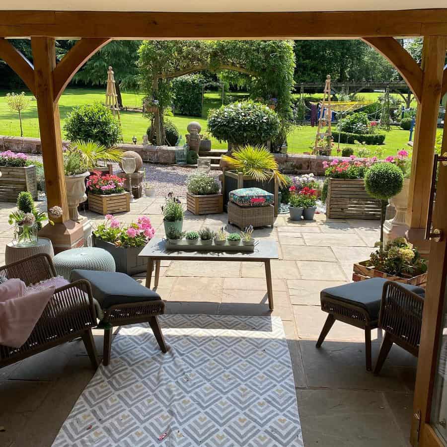 Cozy patio with cushioned chairs and a table under a wooden pergola, overlooking a lush garden with potted plants and colorful flowers