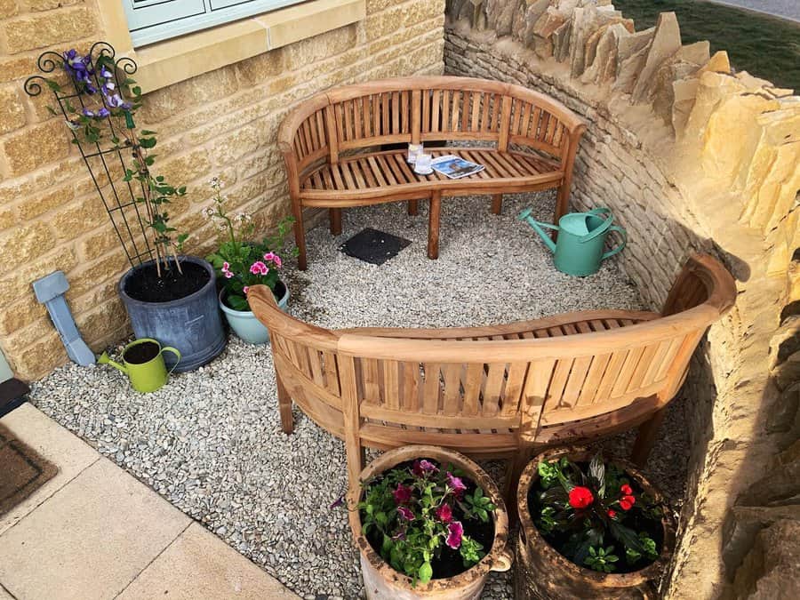 Cozy outdoor nook with two curved wooden benches, potted flowers, a green watering can, and a stone wall backdrop