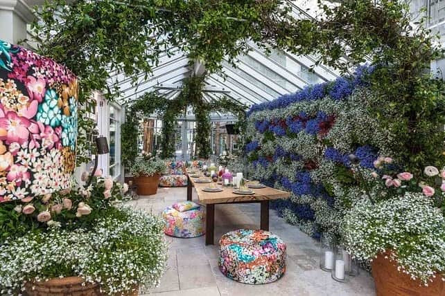 A lush indoor garden with floral cushions, a long wooden table, hanging greenery, and colorful flower-covered walls