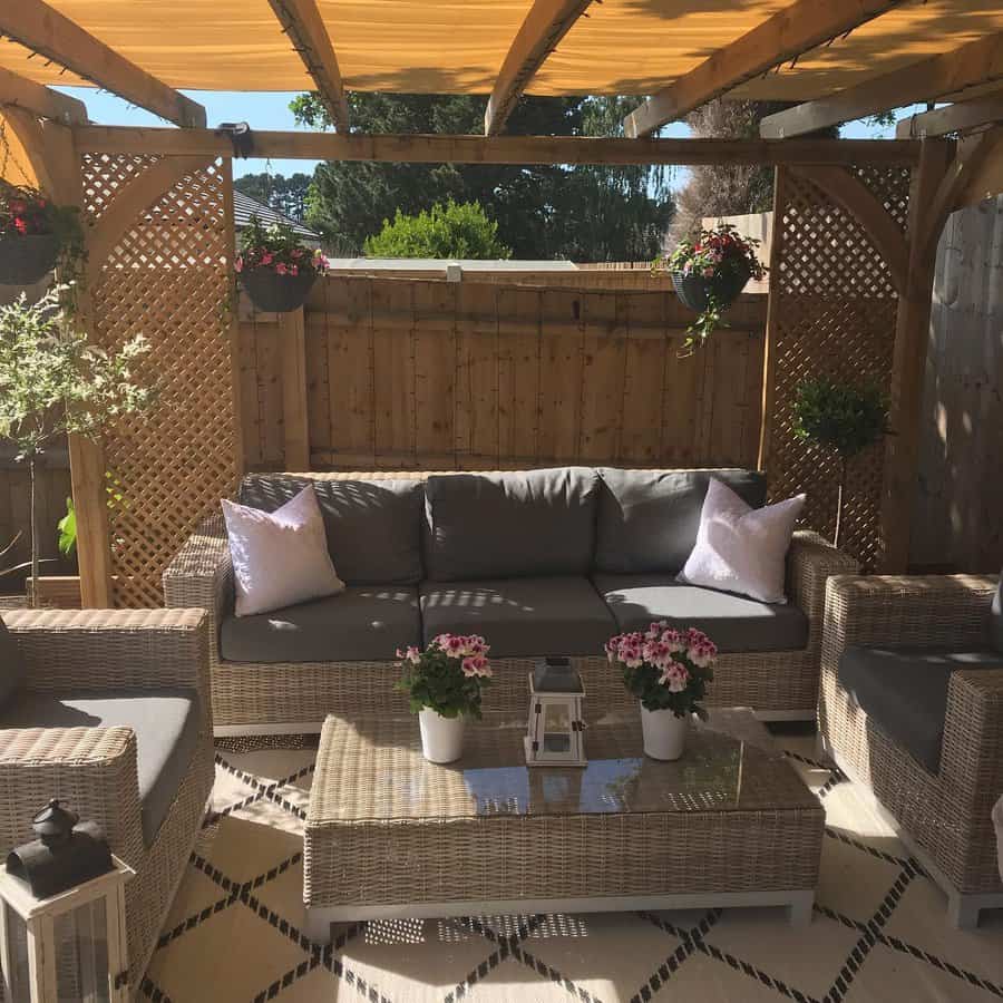 Outdoor patio with wicker furniture, gray cushions, and a pergola. Flowers and lanterns decorate the glass-top coffee table