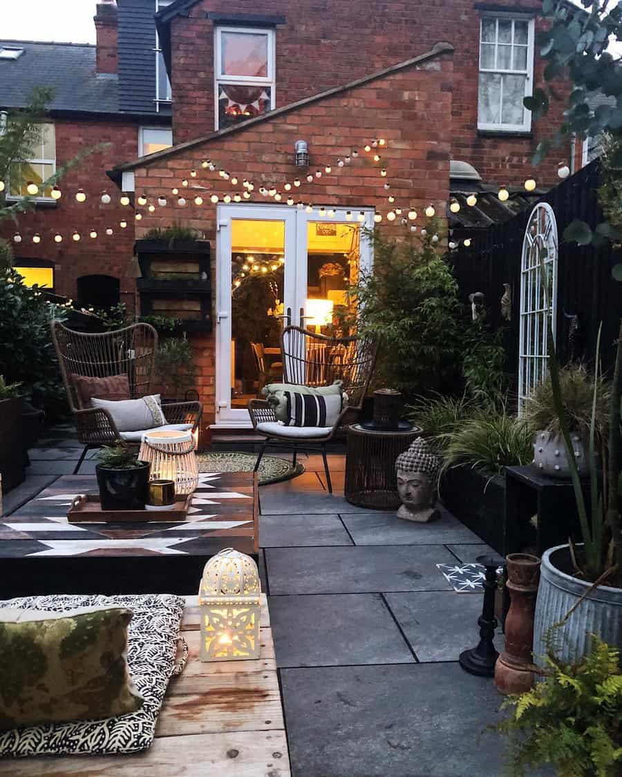 Backyard patio with string lights, wicker chairs, potted plants, and a Buddha head statue, brick house in the background