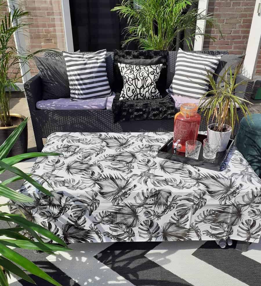 Outdoor patio with a black wicker sofa, tropical-patterned tablecloth, plants, and a tray with a beverage dispenser and cups