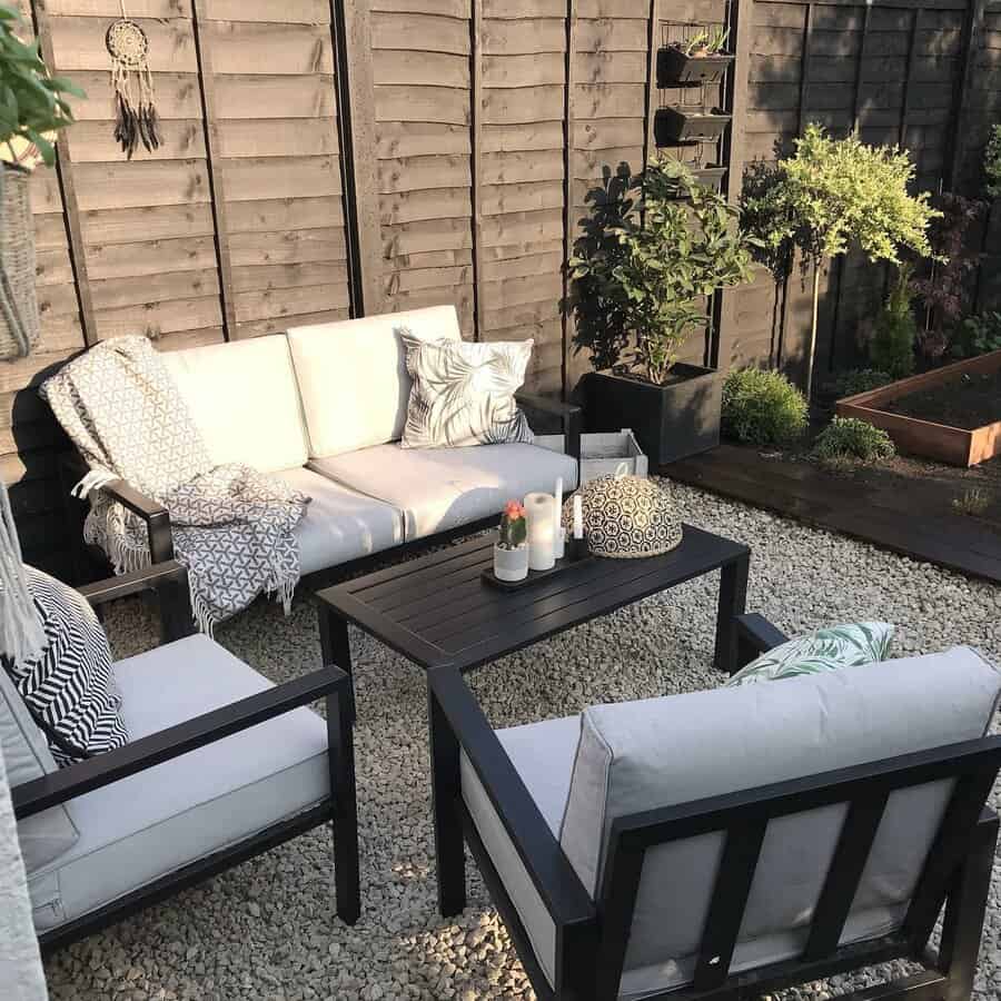 Cozy patio area with white cushioned seating, a dark coffee table, plants, and a wooden fence backdrop