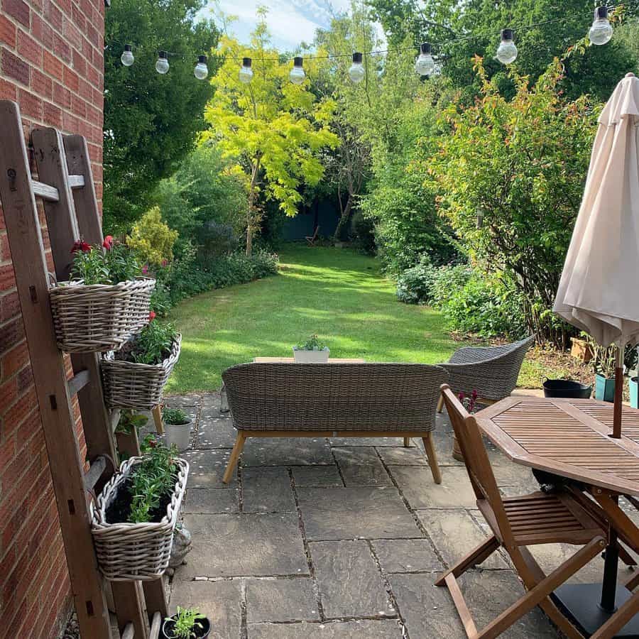 A garden view featuring a wicker sofa, patio chairs, and potted plants under string lights, with a lush green lawn and trees in the background