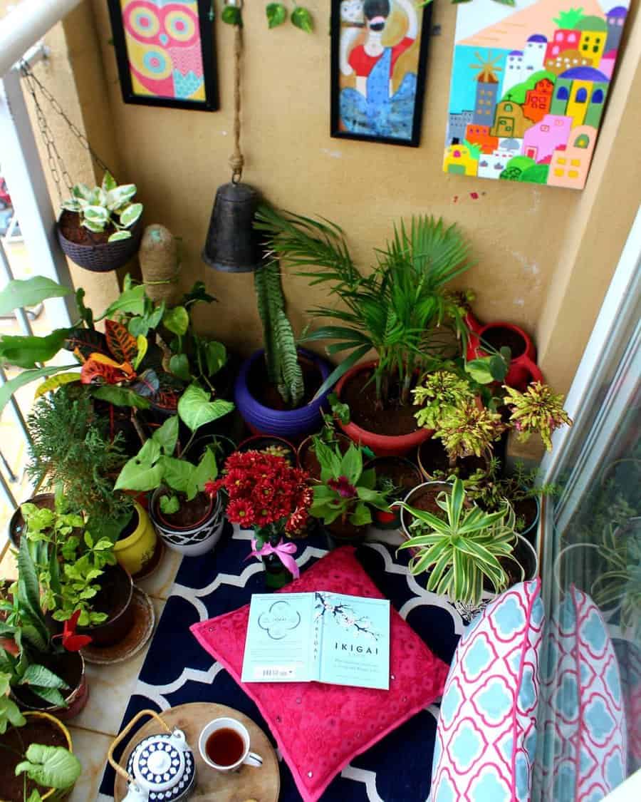 Lush small balcony garden with colorful potted plants, framed artwork, floor seating with cushions, a book, and a tea setup