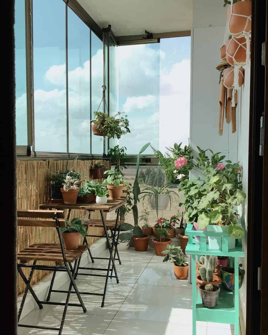 Bright enclosed balcony garden with floor-to-ceiling glass windows, a wooden bistro set, potted greenery, and hanging planters, creating a cozy and airy space