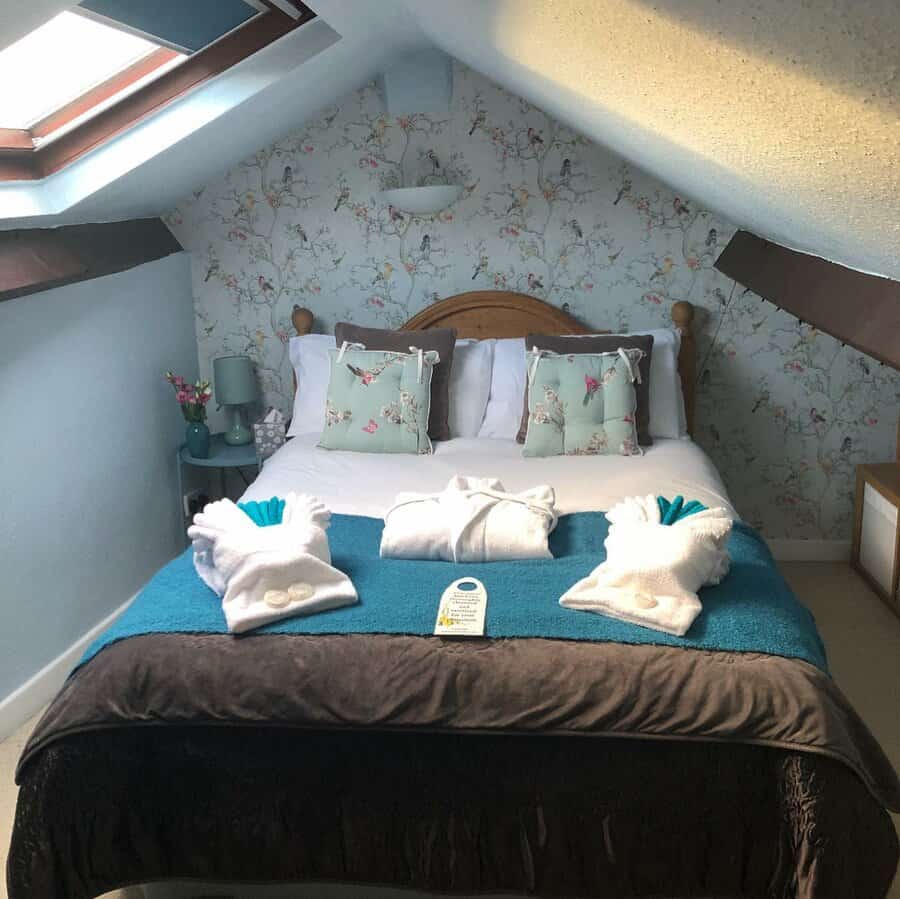 Cozy attic bedroom with floral wallpaper, skylights, and soft blue tones. A warm wooden bed frame and plush bedding create a charming retreat.