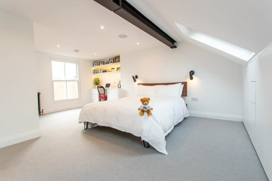 Minimalist attic bedroom with sleek white walls, a skylight, and a cozy workspace. Soft lighting and a teddy bear add warmth to the space.