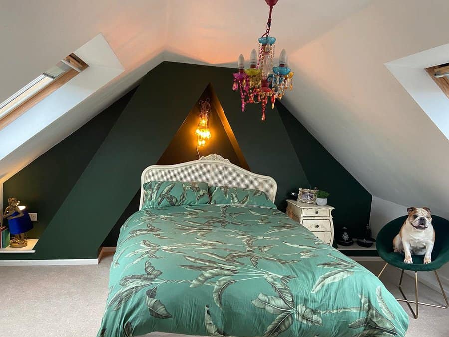 Bold attic bedroom with deep green walls, a statement chandelier, and tropical bedding. Skylights and eclectic decor add a vibrant, cozy feel.