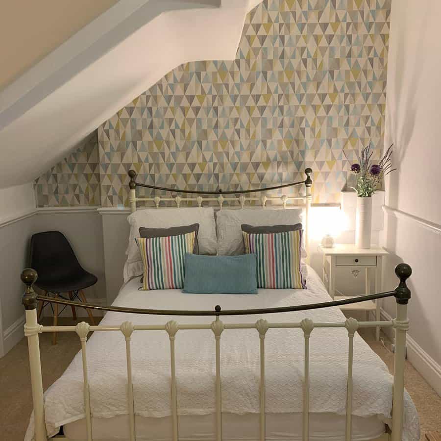 Cozy attic bedroom with a vintage iron bed, geometric wallpaper, and soft lighting. Striped accent pillows add a playful touch to the space.