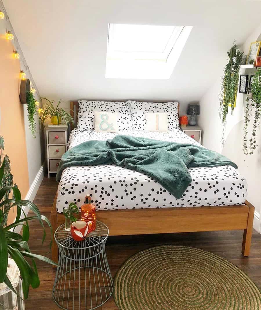 Cozy attic bedroom with polka dot bedding, lush greenery, and warm string lights. A skylight brings in natural light, creating a fresh retreat.