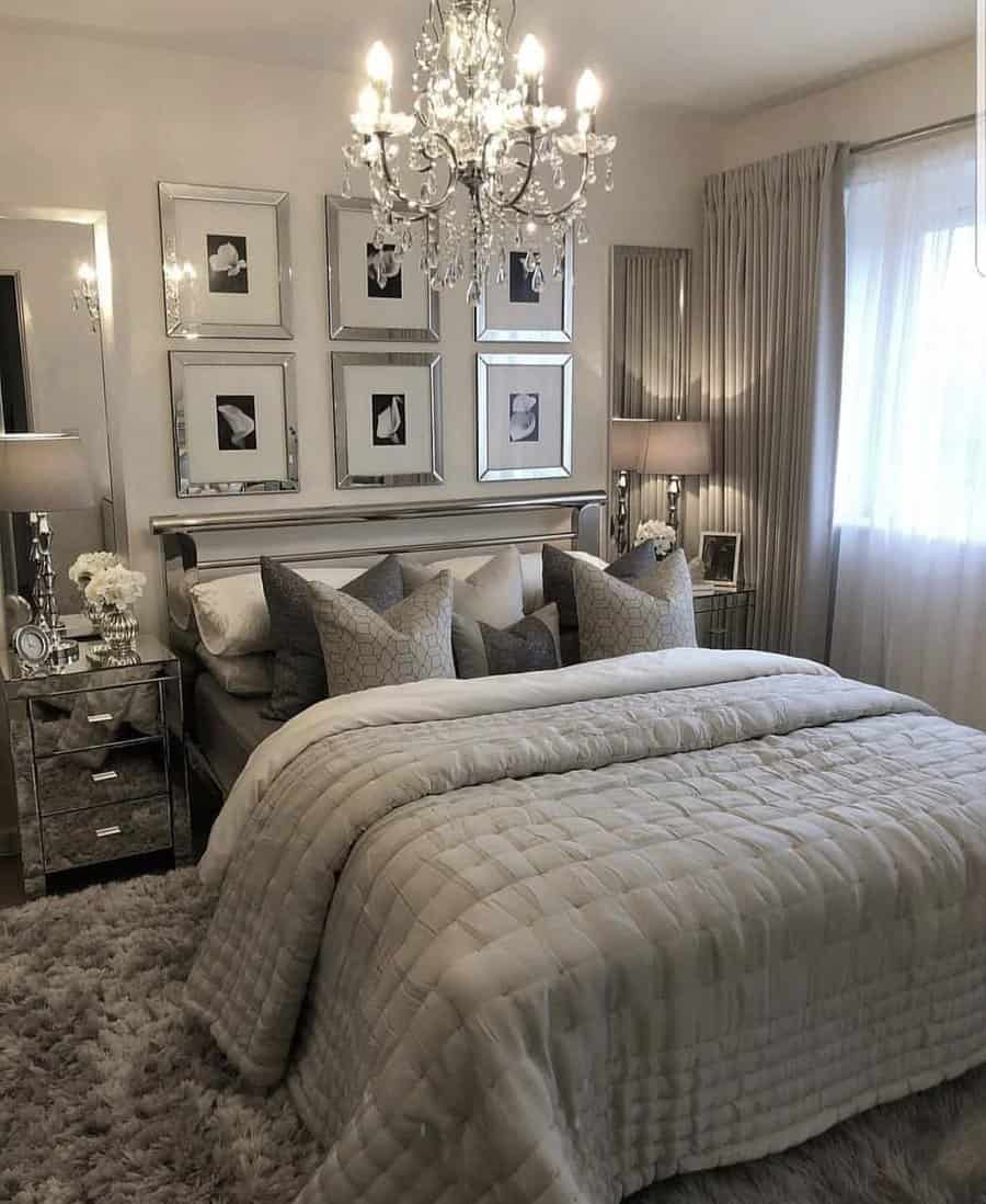 Elegant bedroom with a chandelier, mirrored nightstands, and plush bedding, framed art on walls, and a large window with curtains