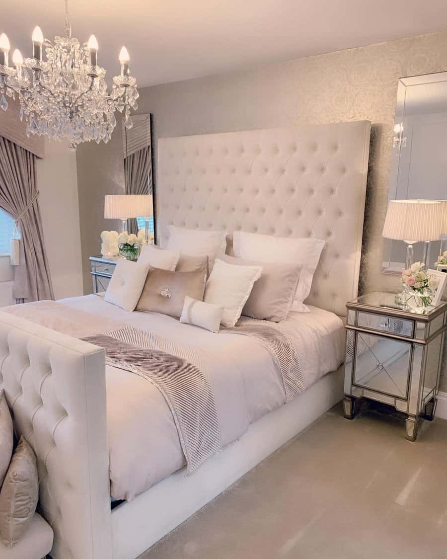 Luxurious bedroom with a tufted headboard bed, mirrored nightstands, elegant lamps, and a crystal chandelier; neutral tones throughout