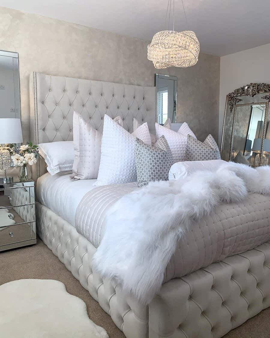 Elegant bedroom with tufted bed, fluffy white bedding, stacked pillows, decorative mirror, nightstand with flowers, and ornate chandelier