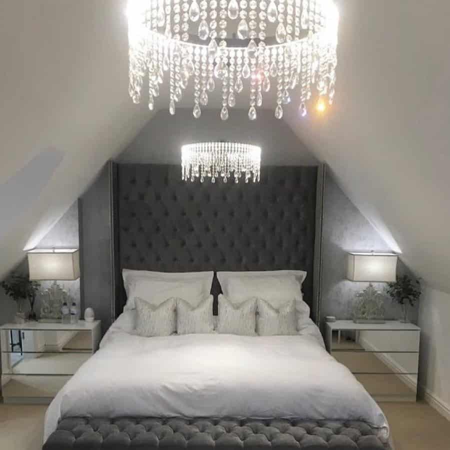 Luxurious attic bedroom with a tufted bed, glass side tables, lamps, and a crystal chandelier; plush decor and soft lighting