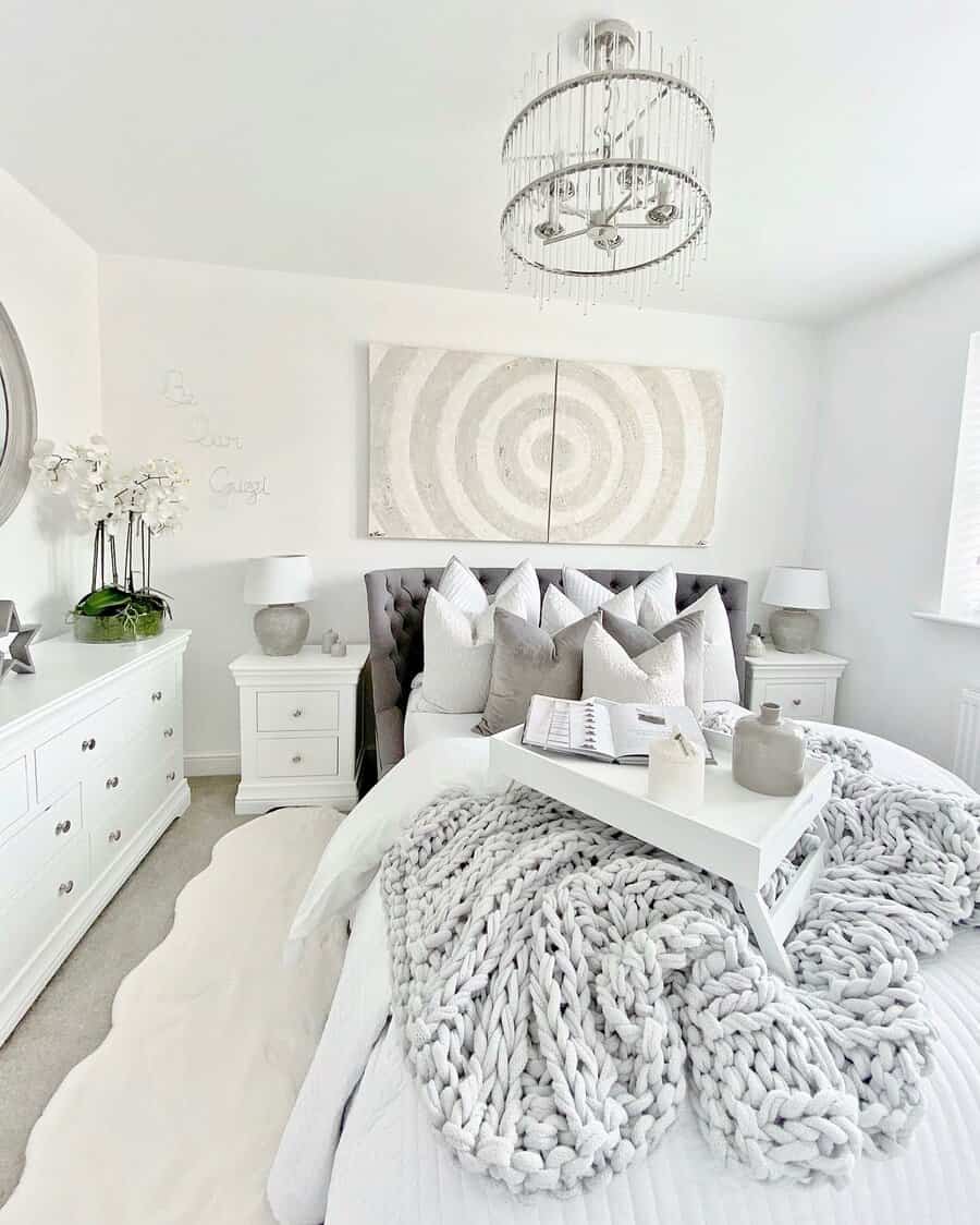 Bright white bedroom with a tufted bed, chunky knit blanket, tray, and modern chandelier. Art decorates the wall above the bed