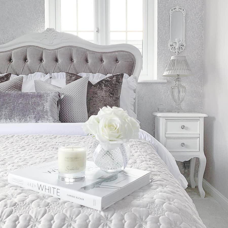 Elegant bedroom featuring a tufted bed, soft pillows, and a bedside table with a vase of white roses and a candle arranged on top of a book