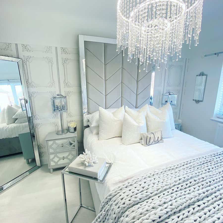 Elegant bedroom with a large mirror, plush white bedding, crystal chandelier, and stylish decor in a soft, neutral palette