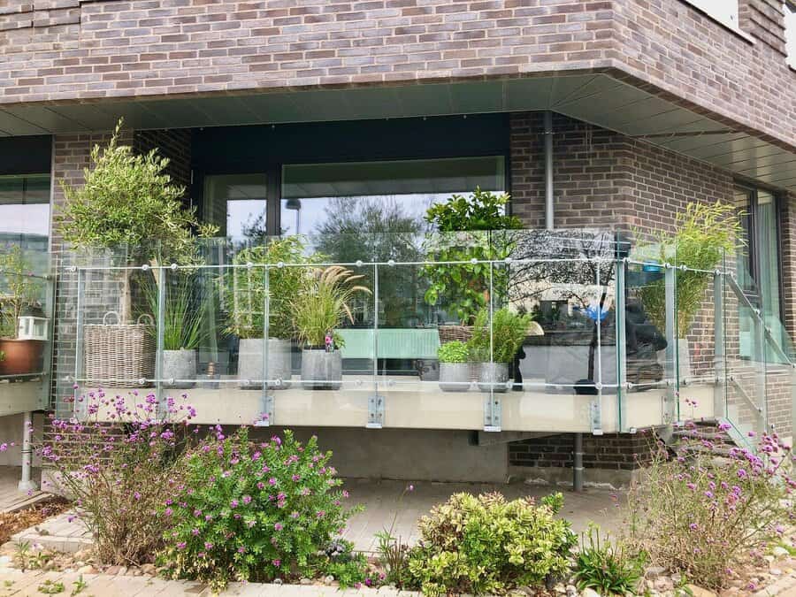 A modern balcony with glass railing, featuring potted plants and a wicker basket, attached to a brick building overlooking a garden