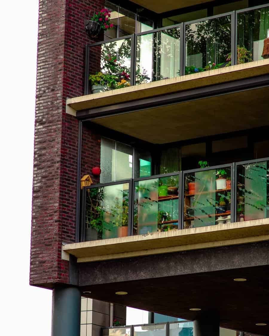 Three apartment balconies with glass railings, decorated with plants and flower pots, brick exterior and a birdhouse visible