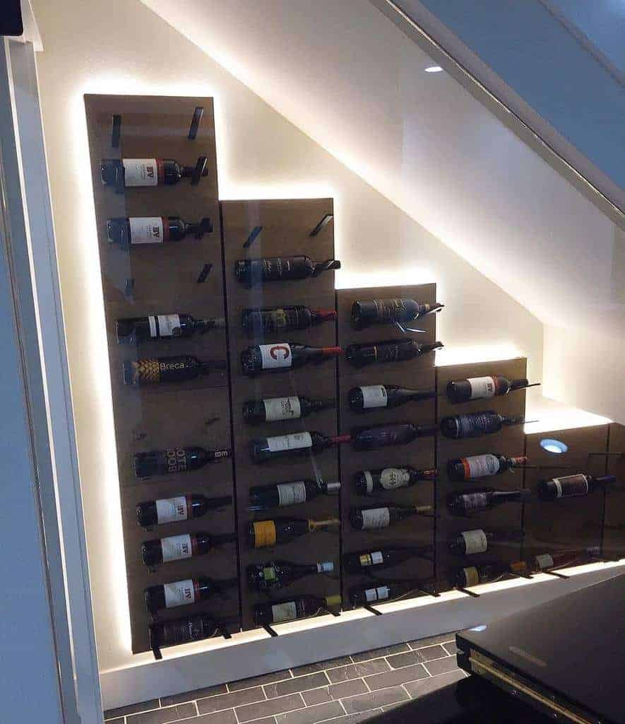 Illuminated wine rack under staircase, bottles displayed horizontally on angled shelves with black tile floor