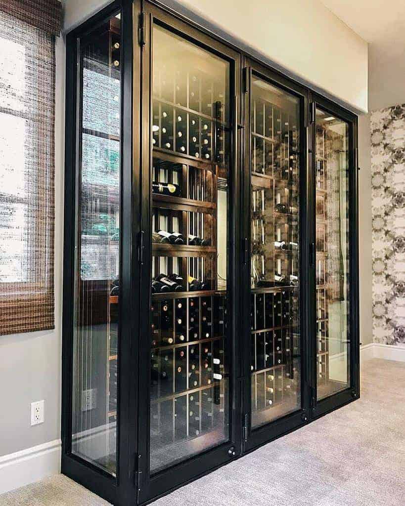 A glass-enclosed wine cellar with black framing, displaying numerous wine bottles on wooden racks in a modern interior