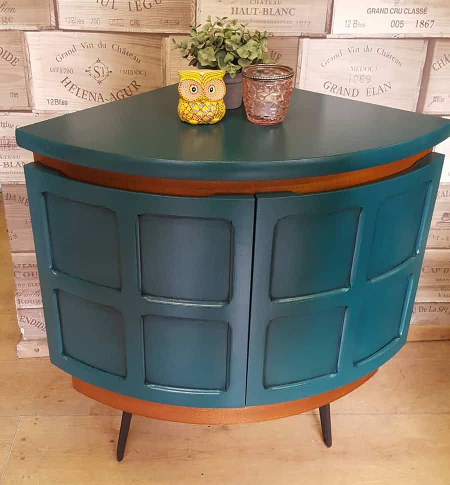 Vintage corner cabinet painted in deep teal with wooden accents and sleek legs, styled with a potted plant and decorative owl figurine.