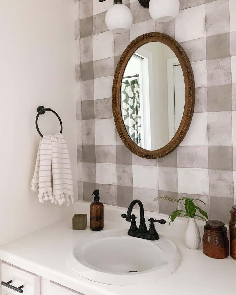 Farmhouse-style bathroom with grey checkered tile backsplash, vintage oval mirror, black faucet, and rustic countertop decor.