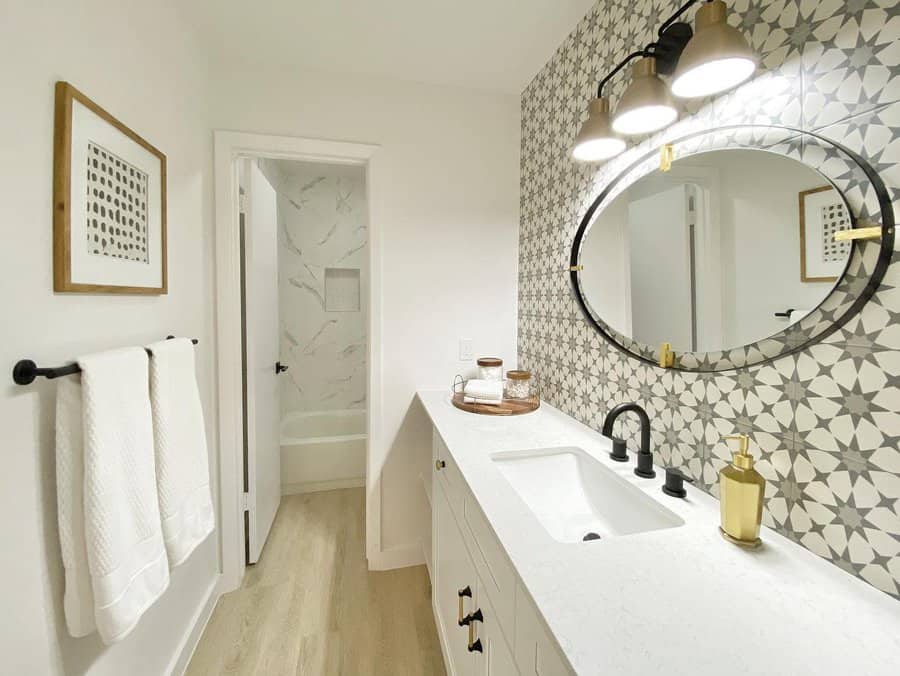 Stylish bathroom with patterned grey tile backsplash, oval mirror with gold accents, black fixtures, and white vanity for a clean contrast.