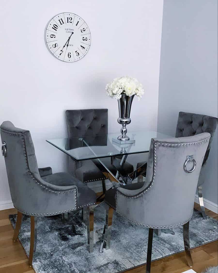 Elegant dining room with a glass table, four gray velvet chairs, a vase of white flowers, and a wall clock above on a light wall