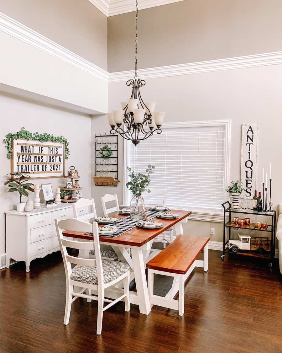 Bright dining room with a wooden table, white chairs, a bench, chandelier, and wall decor including a sign saying "ANTIQUES"