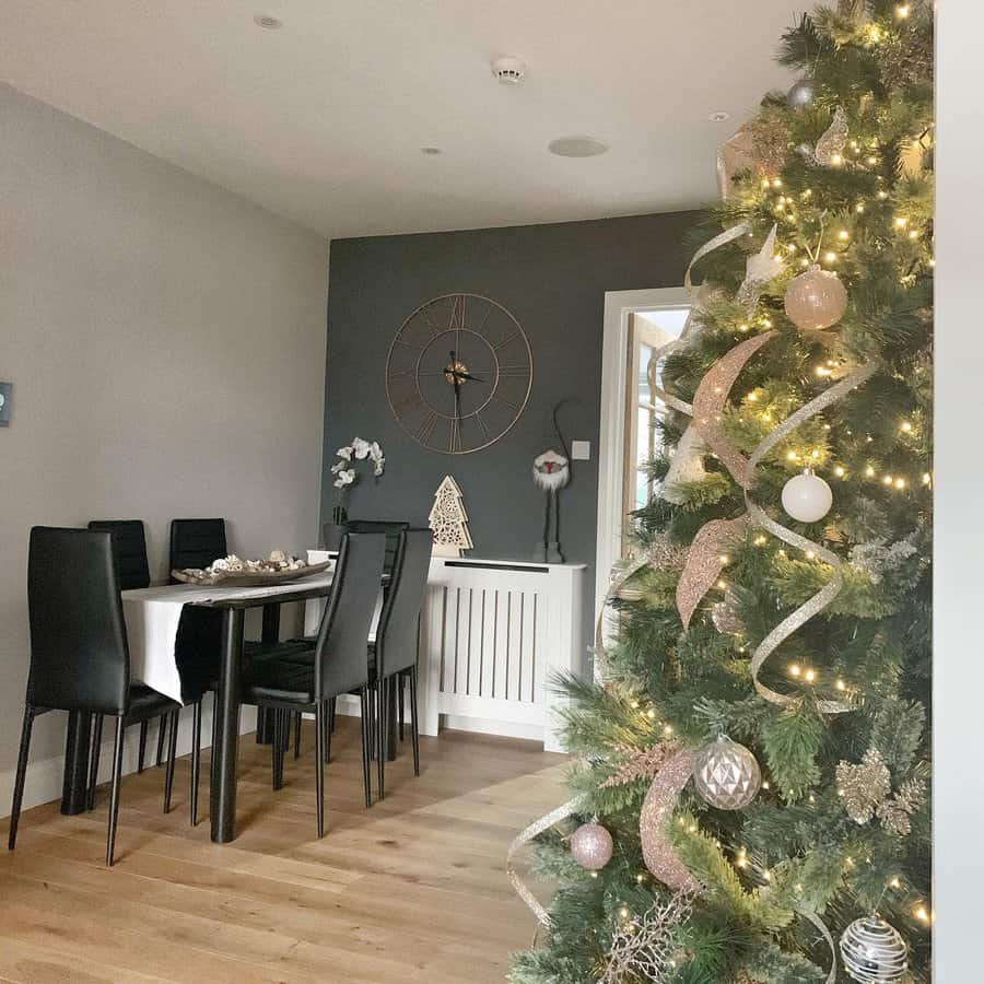 A decorated Christmas tree with lights and ribbons stands in a dining room with a table, chairs, wall clock, and festive decor