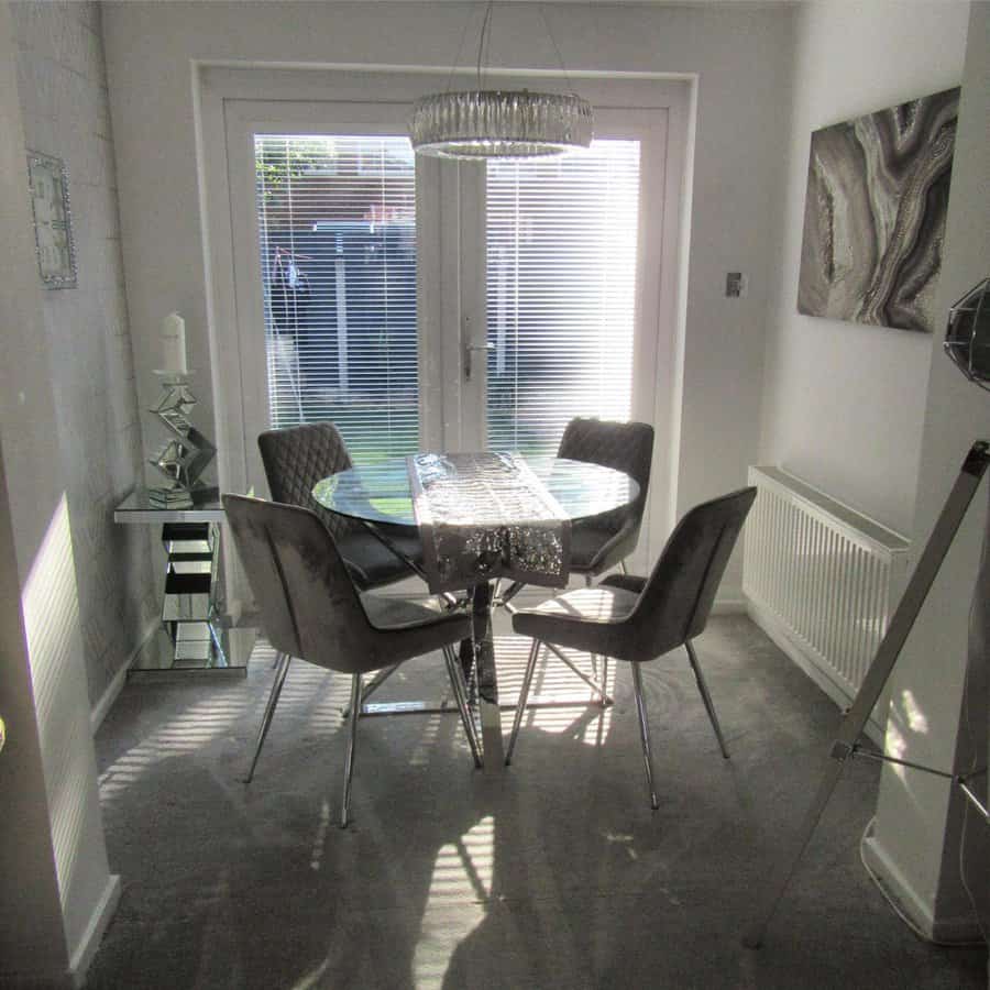 A modern dining area with a round glass table and four gray chairs, a chandelier hangs above, and large windows let in natural light