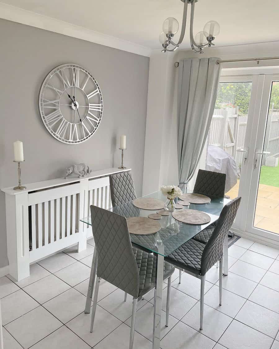 Modern gray dining room with a glass table, four gray quilted chairs, large wall clock, decorative candles, and patio doors to a garden