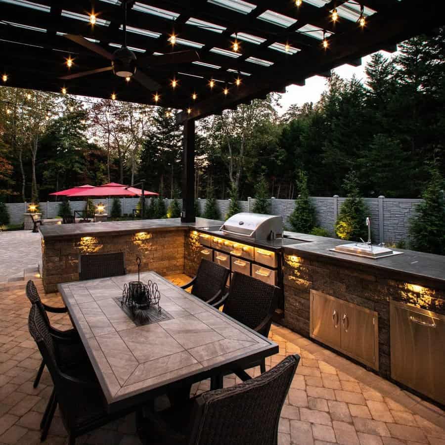 Outdoor patio with a pergola, string lights, dining table, and a built-in kitchen with a grill and sink, trees and umbrellas in the background