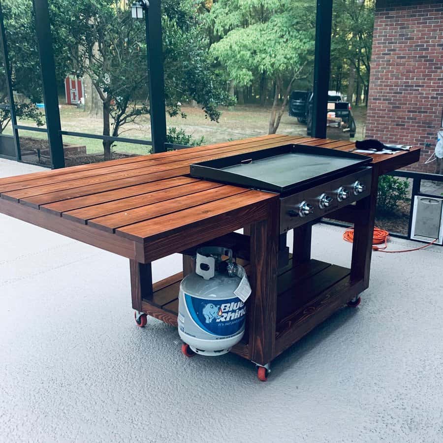 Wooden outdoor grill station with a flat top griddle, propane tank, and shelves, set on a patio with trees in the background