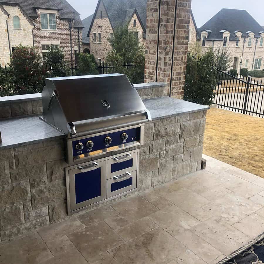 Outdoor kitchen with a built-in stainless steel grill and stone countertops, set on a patio with brick and stone housing in the background