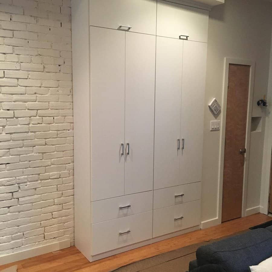 White wardrobe with multiple doors and drawers against a white brick wall in a room with wooden flooring