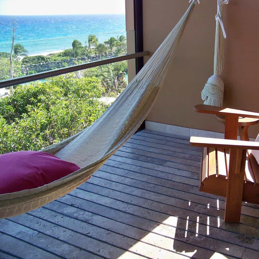 Relaxing balcony with a beige hammock, wooden Adirondack chairs, and an ocean view framed by lush greenery and palm trees