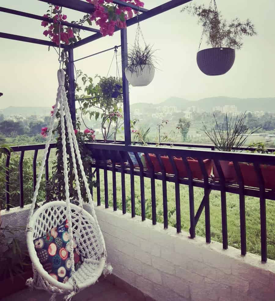 Scenic balcony with a white macramé hanging chair, colorful cushion, hanging planters, and blooming pink flowers, overlooking a lush green landscape and distant city skyline